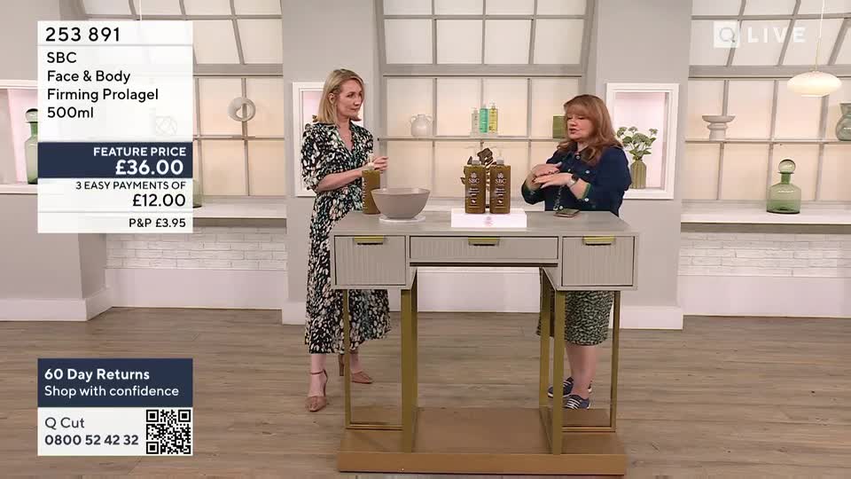 Two women stand behind a desk, one gesturing with her hands as she speaks. The other holds a small glass, listening intently.
