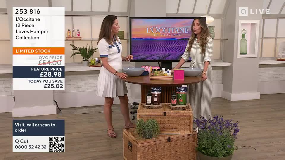 Two women stand beside a round table, showcasing L'Occitane products on a QVC Beauty segment. One woman gestures towards the products, while the other speaks, likely describing the items for sale.
