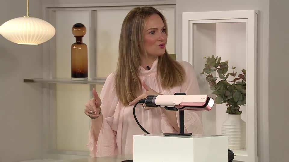 The woman in the pink blouse gestures with her hands, talking about a hair styling tool. A sleek, pink and white flat iron sits on a white pedestal, ready for demonstration.
