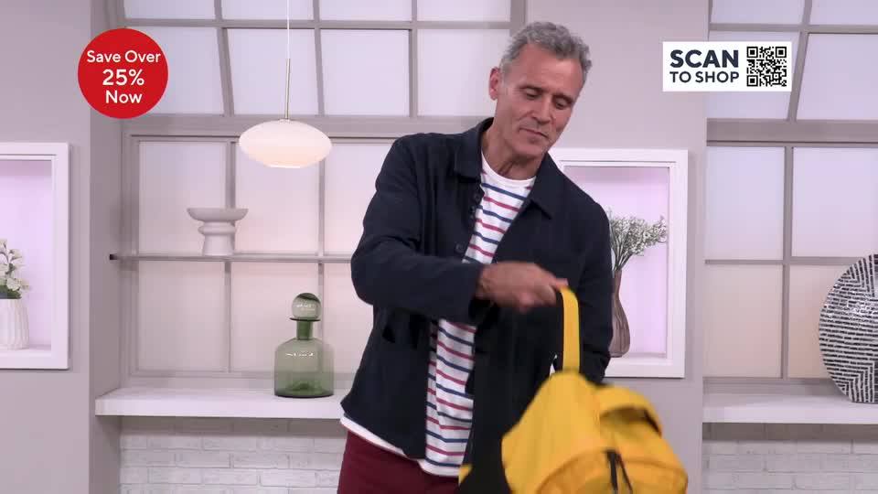 A man in a striped shirt holds up a bright yellow backpack, its straps dangling. He seems to be demonstrating something, perhaps showing off its features on the QVC Beauty set.
