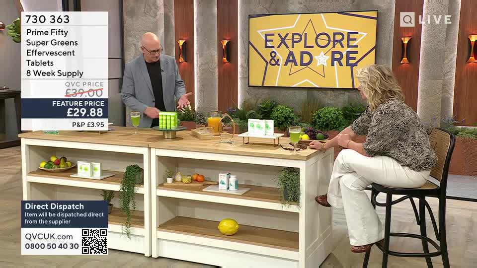 A man in a grey jacket gestures toward a stack of green tablets on a countertop. A woman sits nearby, listening as they present the product on QVC Beauty from the United Kingdom.
