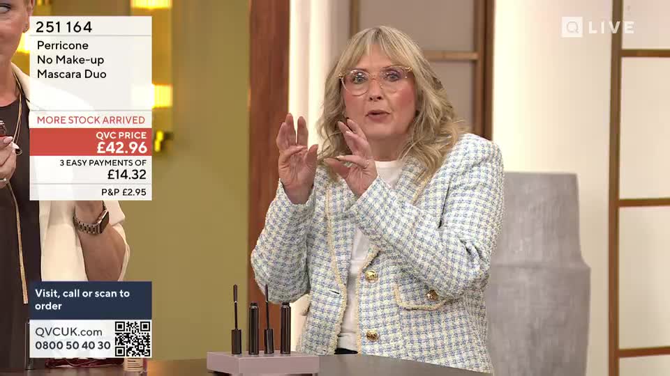 A woman in a tweed jacket gestures with her hands, explaining something about the mascara duo on the table. The QVC Beauty presenter stands in front of a display of the product, ready to demonstrate.
