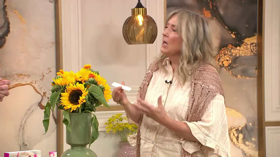 A woman in a cream-colored shirt and shawl gestures with her hands while holding a small white object. Bright yellow sunflowers in a green vase sit nearby, adding a pop of color to the scene.
