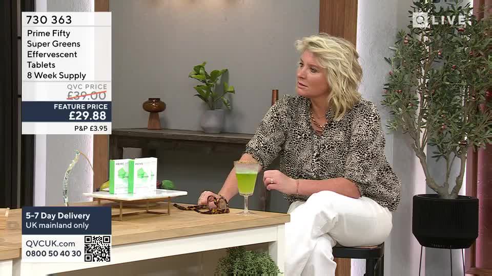 A woman in a patterned blouse sits at a table, gesturing with her right hand. Beside her, a tall glass holds a bright green drink, and a small tray displays boxes of "Super Greens" tablets.
