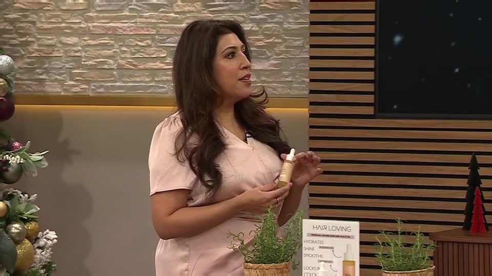 A woman in a pink top holds a small bottle, gesturing with it as she speaks. The set has a Christmas tree and a wooden panel backdrop, typical of a QVC Beauty segment.

