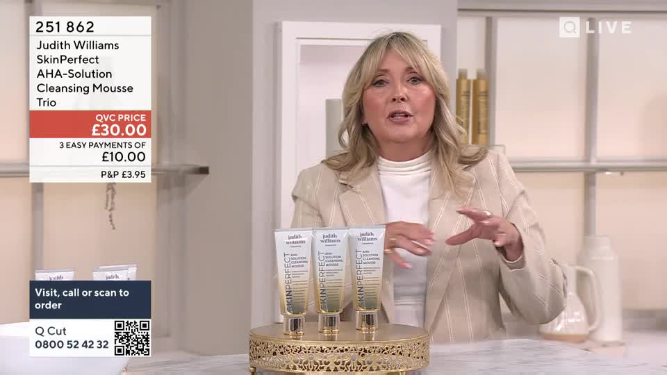 A woman in a cream-colored blazer gestures towards three tubes of skincare on a gold stand. The QVC Beauty presenter is speaking, likely promoting the product to viewers in the United Kingdom.

