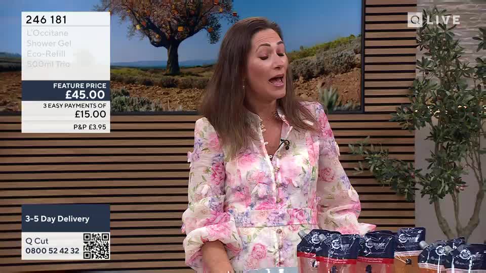 A woman in a floral dress is speaking animatedly, her mouth open as she gestures with her hands. Several packages of what appears to be shower gel sit on the table in front of her, alongside a screen displaying the QVC Beauty product details.
