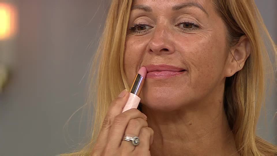 A woman is applying a pale pink lipstick, carefully swiping it across her lower lip. She holds the tube with a ringed hand, her gaze focused on the application.
