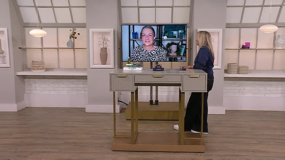 A woman in a dark blue top stands beside a desk, facing a large screen displaying another woman. The presenter is likely in the United Kingdom, as she is on QVC Beauty.
