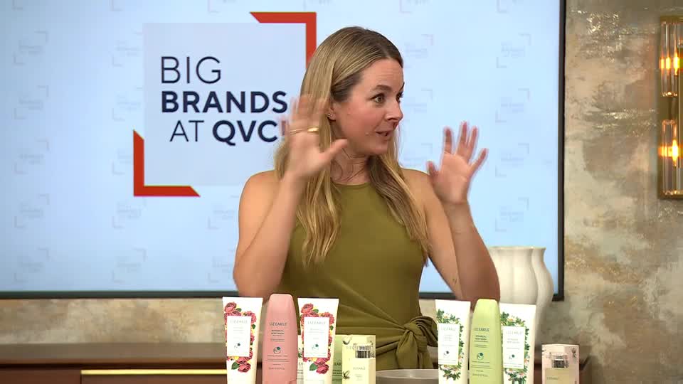A woman in a green dress gestures with both hands, as if explaining something. Behind her, a backdrop reads "BIG BRANDS AT QVC" and various beauty products are arranged on a table.
