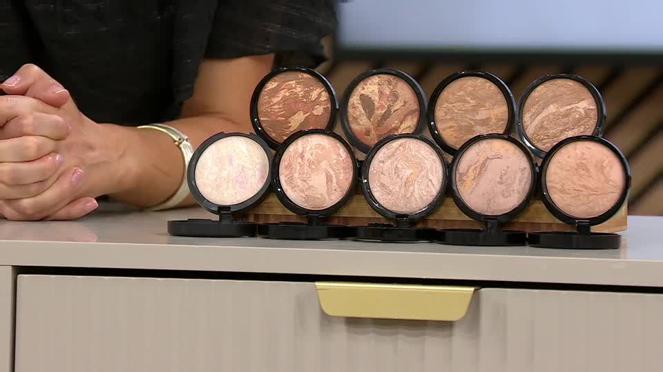 A selection of compact powders sits on a light-colored surface, each with a different marbled pattern. The presenter's hands are clasped in front of them, ready to demonstrate the products.
