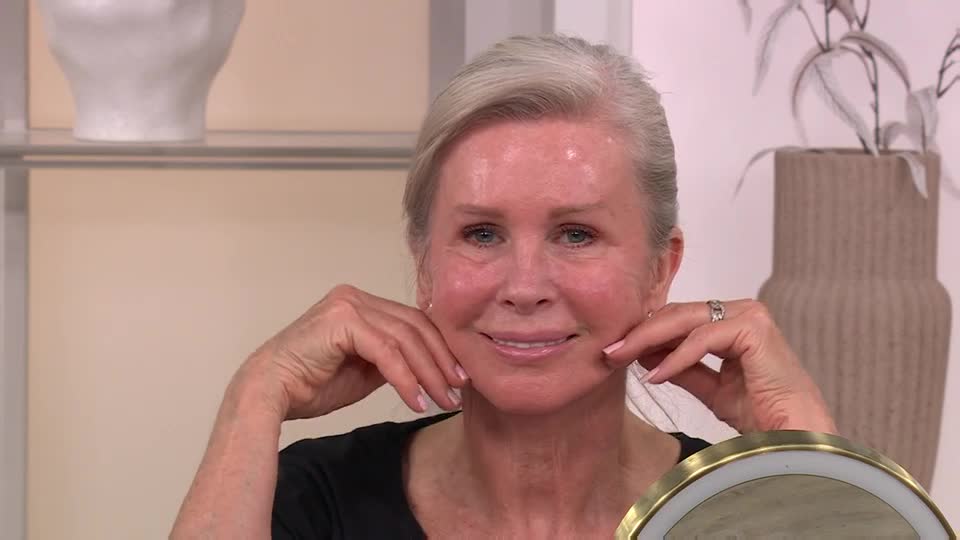 A woman with silver hair gently pinches her cheeks, smiling at the camera. She's in a brightly lit studio, likely demonstrating a beauty product on QVC UK.
