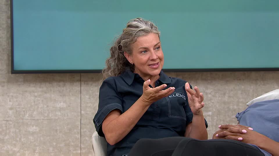 The woman in the dark shirt gestures with her hands, explaining something to someone off-camera. The backdrop is a muted teal, and the logo on her shirt reads "ELEMIS."
