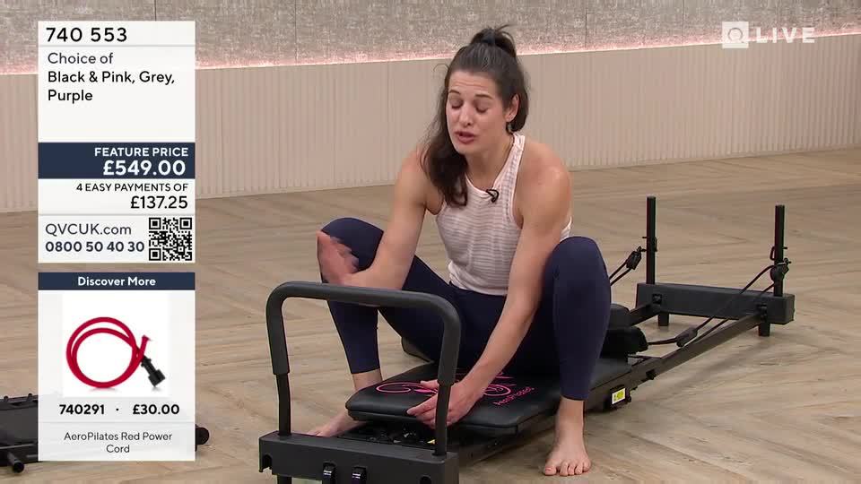 A woman sits on a Pilates machine, demonstrating its use. She's positioned low, her legs bent, and her hands are on the platform.
