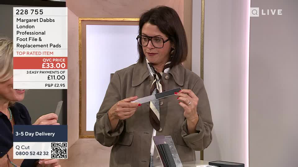 A woman in a blazer holds a foot file, turning it slightly to show its features. The QVC Beauty presenter gestures with the foot file, presumably demonstrating its use.
