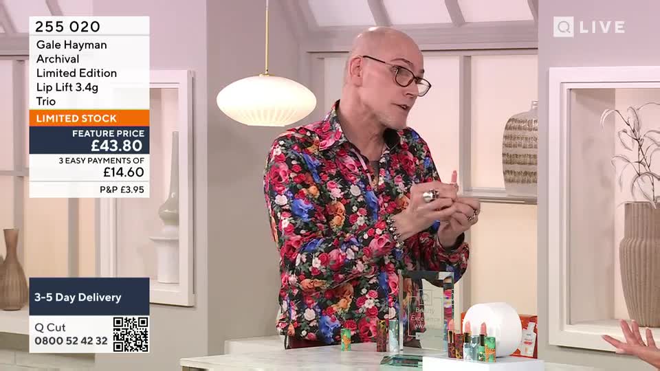 The man in the floral shirt gestures with his hands while talking, holding what looks like a lipstick. A display of lipsticks and other beauty products sits on the table in front of him.
