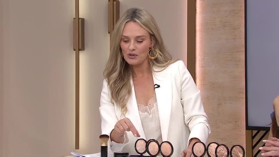 The woman in the white blazer gestures toward a row of compacts, her finger pointing at one. She's on a set, likely for a QVC Beauty segment, and talking to someone off-camera.
