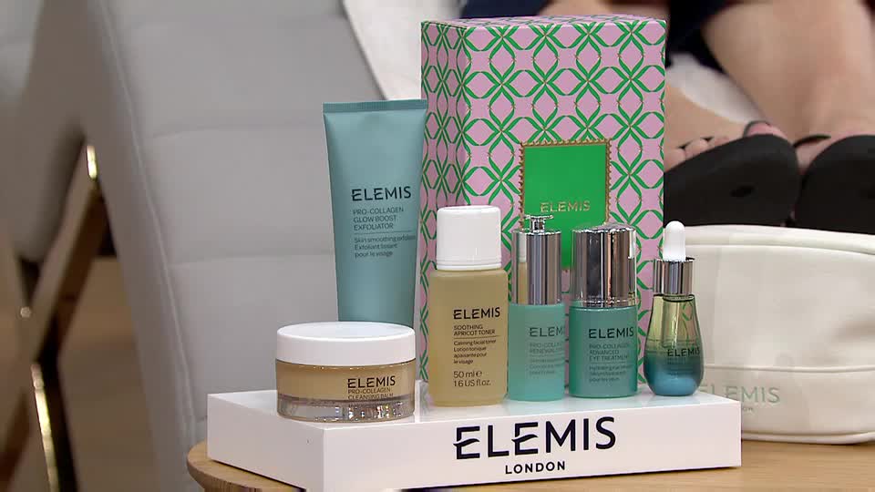 A collection of Elemis skincare products is arranged on a white platform, with a patterned box and a makeup bag in the background. The presenter's feet, clad in black sandals, are visible near the top of the frame.
