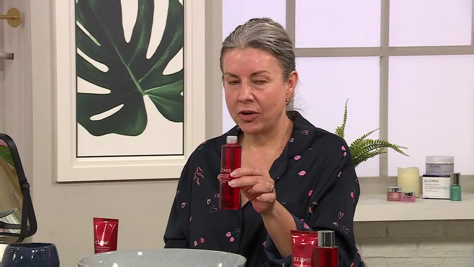 A woman in a dark floral top holds a red bottle, speaking directly to the camera. Behind her, a framed botanical print hangs on the wall, and beauty products are arranged on a nearby table.
