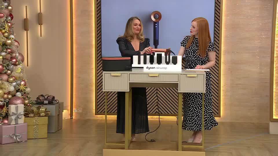 Two women stand at a small desk, discussing a Dyson Airwrap. A Christmas tree and wrapped gifts decorate the set, suggesting a QVC Beauty segment from the United Kingdom.
Two women stand at a small desk, discussing a Dyson Airwrap. A Christmas tree and wrapped gifts decorate the set, suggesting a QVC Beauty segment from the United Kingdom.
