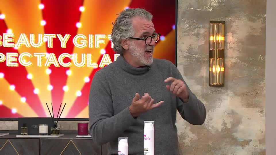 A man in a grey turtleneck gestures with his hands, speaking passionately. He stands in front of a screen advertising a "Beauty Gift Spectacular" on QVC, with two white bottles of skincare products on a nearby table.
A man in a grey turtleneck gestures with his hands, speaking passionately. He stands in front of a screen advertising a "Beauty Gift Spectacular" on QVC, with two white bottles of skincare products on a nearby table.