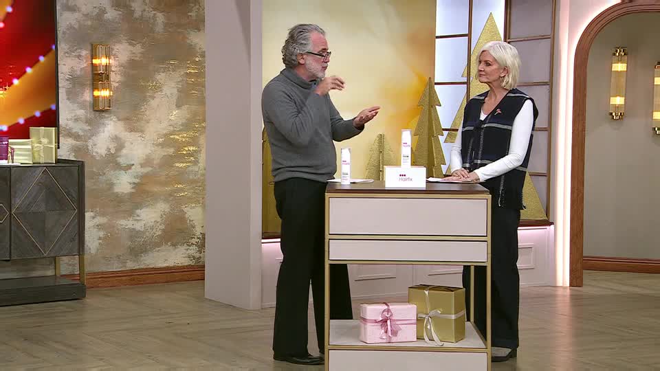 The man gestures towards the skincare products on the table, while the woman stands beside him, looking on. The set is decorated with gold and pink gift boxes, suggesting a QVC Beauty segment from the UK.
The man gestures towards the skincare products on the table, while the woman stands beside him, looking on. The set is decorated with gold and pink gift boxes, suggesting a QVC Beauty segment from the UK.