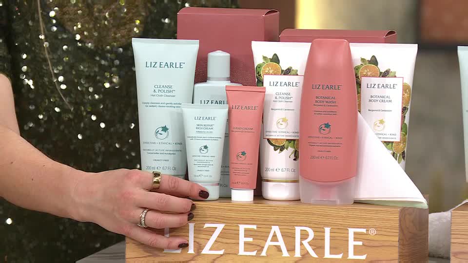 A hand, adorned with rings, arranges a collection of Liz Earle skincare products. The bottles and tubes, in varying shades of white and pink, are displayed on a wooden platform bearing the brand's name.
