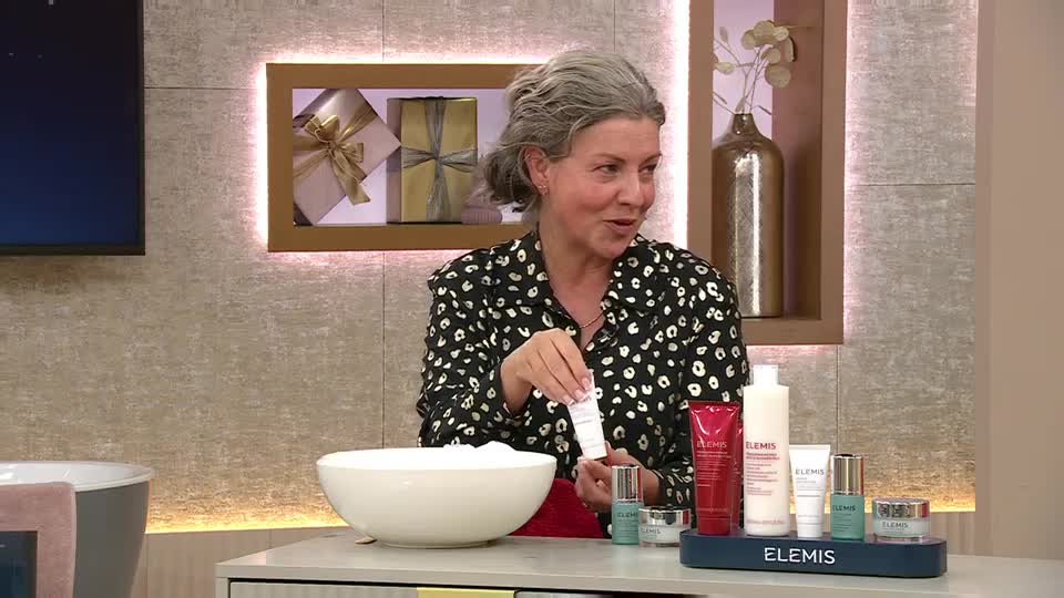 A woman in a black and white patterned shirt holds a small white tube, gesturing toward a collection of Elemis products. She's in a studio setting, likely a QVC Beauty segment from the United Kingdom, with a large bowl and a bathtub nearby.
