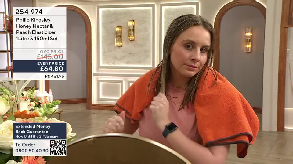 A woman, fresh out of the shower, holds an orange towel around her shoulders while she examines her wet hair. She's on a QVC Beauty segment, likely in the United Kingdom, and the product details are displayed on a screen behind her.
