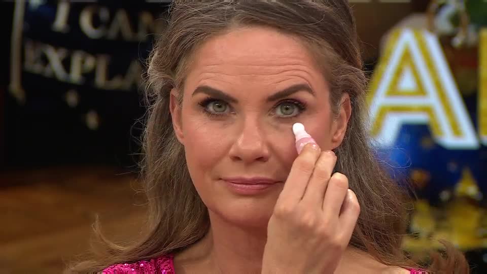 On a QVC Beauty segment from the United Kingdom, a woman is gently dabbing a small, pink-and-white applicator under her eye. She looks directly at the camera, her expression calm, as she demonstrates the product.
