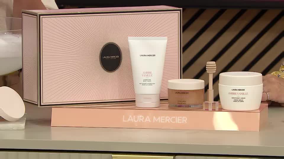 A Laura Mercier beauty set, with its signature packaging, is arranged on a display stand. The presenter's hand is visible, likely about to demonstrate the product on the QVC Beauty show.
