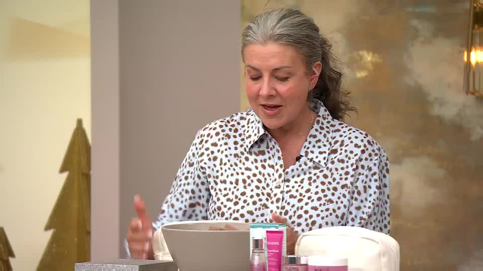 A woman with grey hair pulled back in a ponytail gestures with her hands while speaking. She's wearing a patterned shirt and sits behind a table with various beauty products.