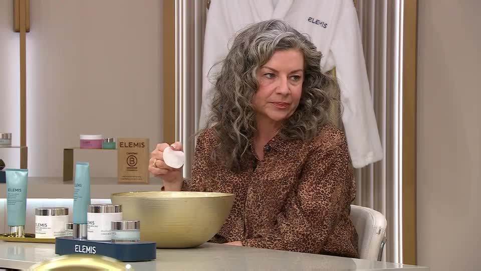 A woman holds a cotton pad over a golden bowl, surrounded by Elemis skincare products. She's on QVC Beauty, demonstrating a routine.