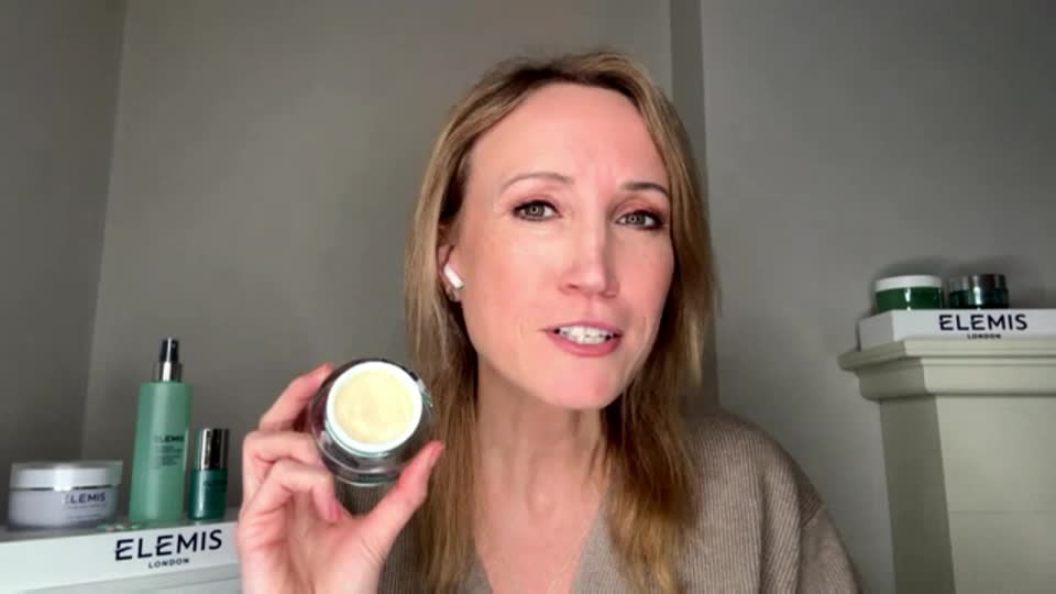 A woman holds up a jar of cream, its pale yellow contents visible. Behind her, several Elemis products sit on a shelf, suggesting a beauty demonstration.