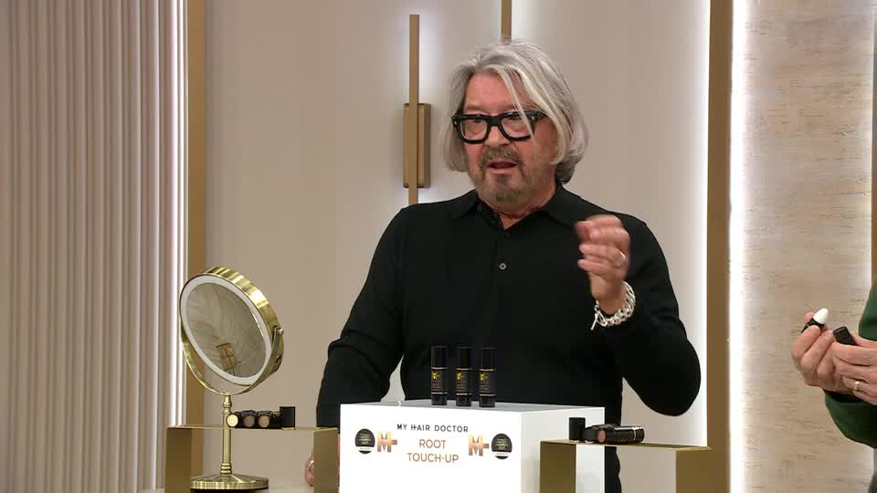 A man with grey hair and glasses gestures with his right hand, explaining something. On a display in front of him are three small bottles of "My Hair Doctor Root Touch-Up."