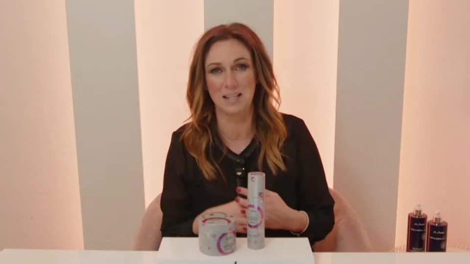 A woman with auburn hair speaks directly to the camera, her hands resting on a white surface. Beside her, a tall, silver bottle with pink accents stands upright.