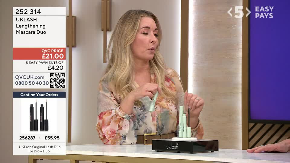 A woman on QVC UK holds up a pale green mascara tube, demonstrating its application.  Several other tubes stand upright on a black display.