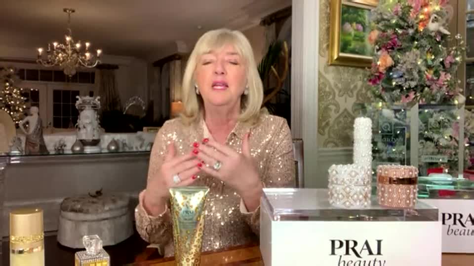 A woman in a sparkly top gestures animatedly, holding a gold and teal bottle of PRAI Beauty. Behind her, a festive Christmas tree twinkles in a room that looks like it's set up for a QVC broadcast.