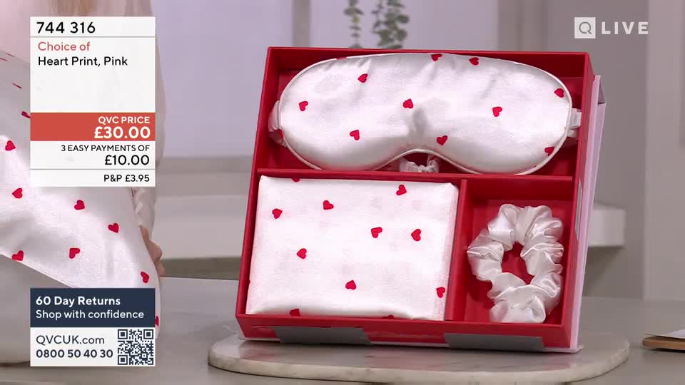 A white silk eye mask and scrunchie, both adorned with tiny red hearts, sit in a red gift box. Beside them, a folded white silk scarf, also patterned with red hearts, rests neatly.