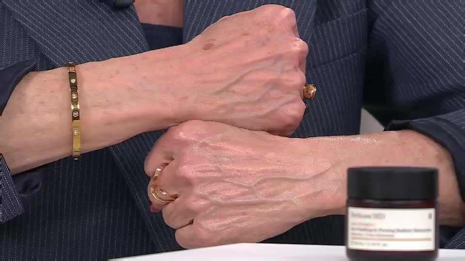 A woman's hands, adorned with gold rings and a diamond bracelet, are held up, showcasing their skin. A small jar of skincare sits nearby on a white surface.