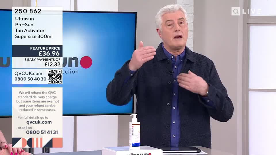 A man in a dark jacket gestures with his hands as he speaks. A screen behind him displays product information for Ultrasun Tan Activator.