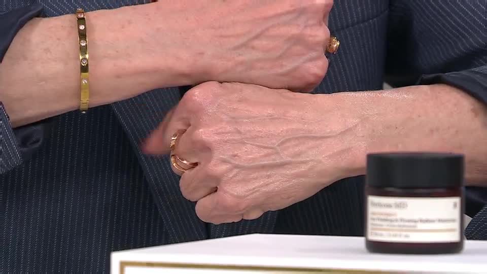 A woman's hands, adorned with a gold bracelet and rings, are positioned near a small jar of skincare. The veins on her skin are clearly visible as she gestures.