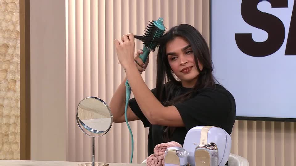 A woman is demonstrating a hair styling tool, its bristles spinning as she guides it through her dark hair. Beside her, a vanity mirror reflects the soft lighting of the studio. A woman is demonstrating a hair styling tool, its bristles spinning as she guides it through her dark hair. Beside her, a vanity mirror reflects the soft lighting of the studio.