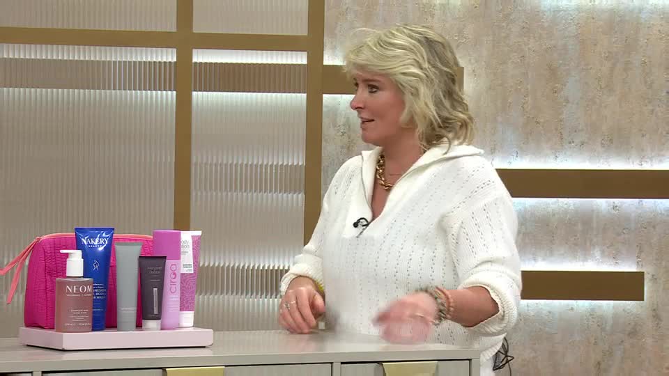 A woman in a white sweater gestures towards a collection of beauty products arranged on a pink tray. She's on a QVC broadcast, likely explaining the benefits of these items. A woman in a white sweater gestures towards a collection of beauty products arranged on a pink tray. She's on a QVC broadcast, likely explaining the benefits of these items.
