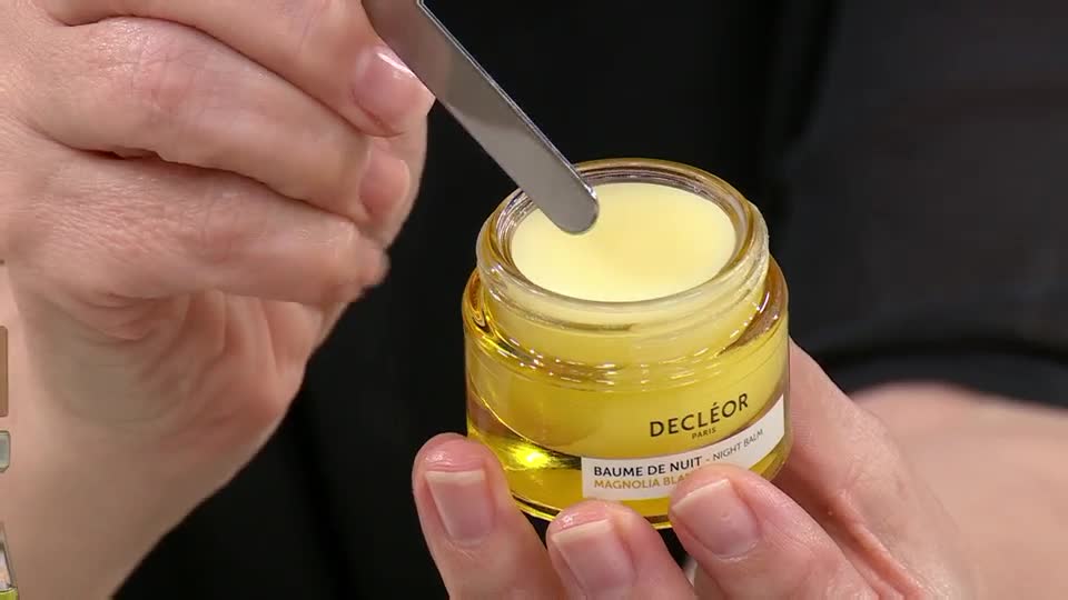 A small metal spatula scoops a dollop of pale yellow balm from a glass jar. The jar, labelled "DECLÉOR PARIS BAUME DE NUIT - NIGHT BALM MAGNOLIA BLANC," is held securely by a hand. A small metal spatula scoops a dollop of pale yellow balm from a glass jar. The jar, labelled "DECLÉOR PARIS BAUME DE NUIT - NIGHT BALM MAGNOLIA BLANC," is held securely by a hand.