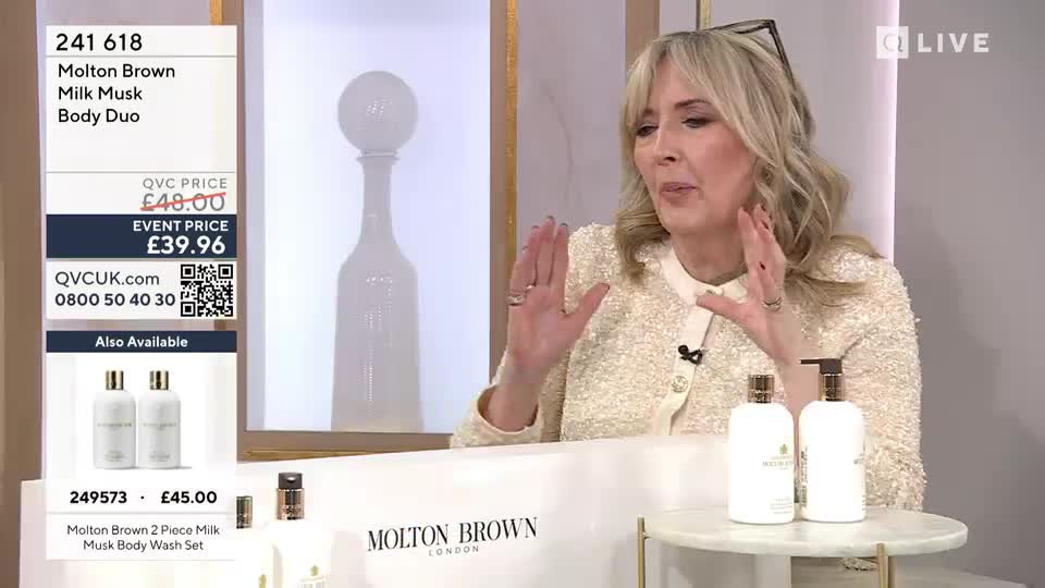 A woman on QVC UK is enthusiastically describing a Molton Brown Milk Musk Body Duo. Two elegant white bottles of the product sit on a small marble-topped table in front of her. A woman on QVC UK is enthusiastically describing a Molton Brown Milk Musk Body Duo. Two elegant white bottles of the product sit on a small marble-topped table in front of her.