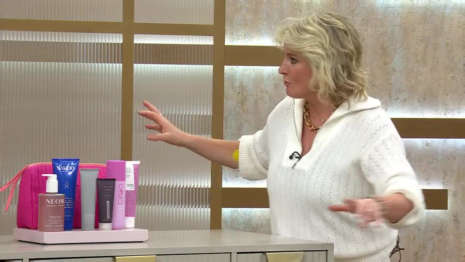 A woman in a white knitted jumper gestures towards a collection of beauty products. The items, including a pink bag and several bottles and tubes, are neatly arranged on a pale pink tray. A woman in a white knitted jumper gestures towards a collection of beauty products. The items, including a pink bag and several bottles and tubes, are neatly arranged on a pale pink tray.