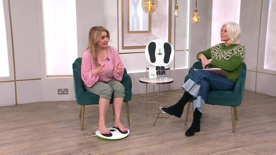 A woman in a pink top stands on a white and green foot massager, talking animatedly to another woman seated in a green armchair. The second woman, wearing a patterned green jumper and wide-cuffed jeans, listens attentively while holding a tablet.