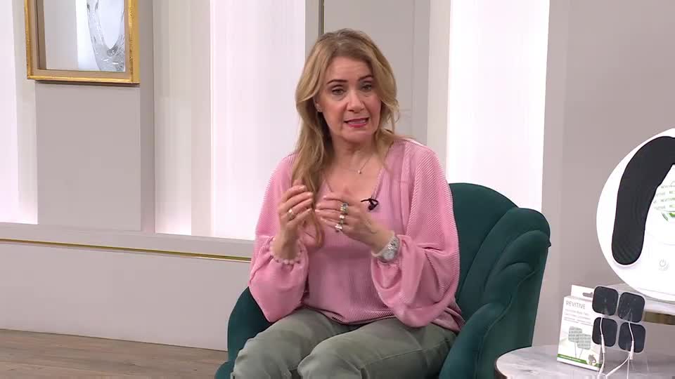 A woman in a pink top gestures with her hands while sitting in a plush chair. On a table beside her, a white device with a black curved pad and several small electrode pads are displayed.