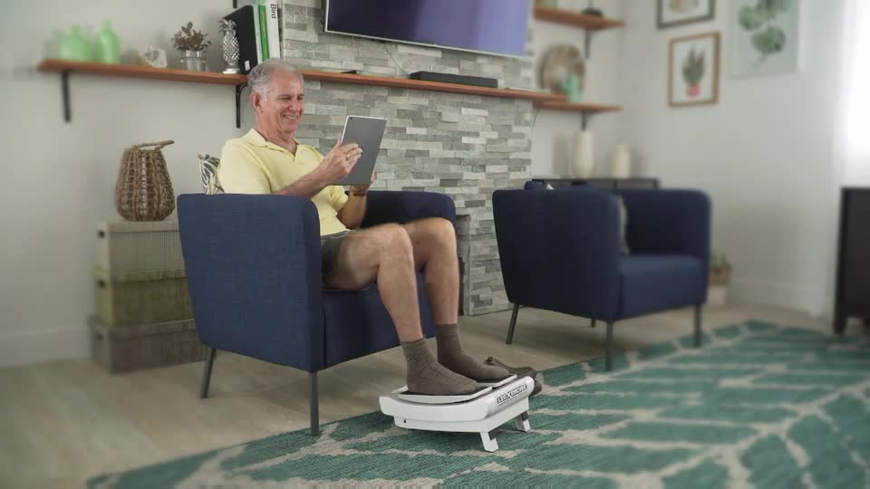 A man sits in a navy armchair, engrossed in his tablet while his feet rest on a white foot massager. The device hums gently, its rollers working beneath his grey socks.